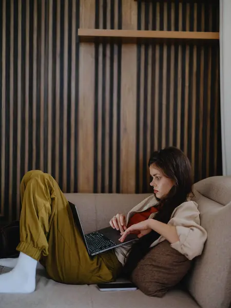 A woman lies on a couch with a laptop and stares pensively into the screen training a freelancer at work, a real lifestyle without retouching. High quality photo