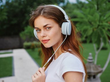 Beyaz tişörtlü, kulaklıklı kadın portresi müzik dinliyor ve sokakta yürüyor, gülümsüyor, arkasında palmiye ağaçları var. Yüksek kalite fotoğraf