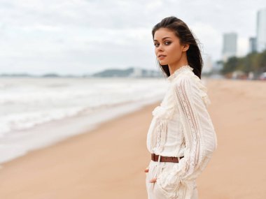 Deniz kenarındaki yaz güzeli: Güzel Beyaz Elbiseli Genç bayan model, Tropikal bir plajda duruyor, Çekici Günbatımı Skyline altında rahatlamanın tadını çıkarıyor..