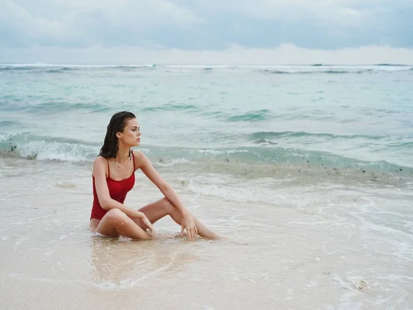A woman with a tanned body and long, wet hair in a red swimsuit sits on the sand by the ocean in the waves on Bali Island Beach and looks out at the beautiful view. High quality photo