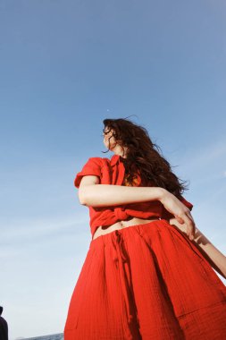 Deniz kenarındaki neşeli dans: Gülümseyen bir kadın, güzel sahil ve mavi gökyüzü ile çevrili, Moda Kırmızı Yazda Özgürlük ve Eğlenceyi Kucakladı