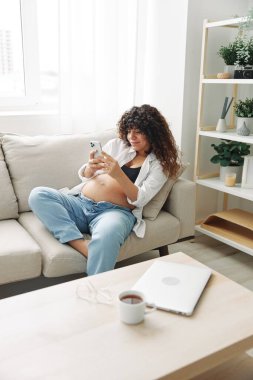 Hamile kadın blogcu evdeki kanepede oturur ve telefonda kendi fotoğraflarını çeker, selfie ve video görüşmesi, doktorla online istişare, hamilelik yönetimi. Yüksek kalite fotoğraf
