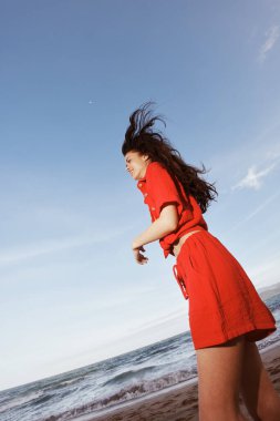 Yaz melteminde dans eden neşeli kadın, şık kırmızı elbiseler giyiyor, plaj tatilinin keyfini çıkarıyor.