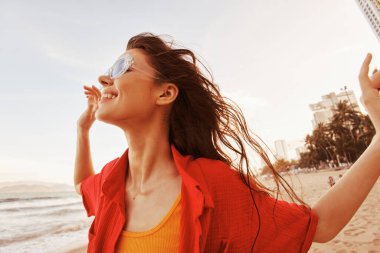 Güneş Gözlüklü Gülümseyen Kadın, Özgürlüğü Kucaklayan: Sahilde Günbatımında Neşeli Güzelliğin Canlı Portresi