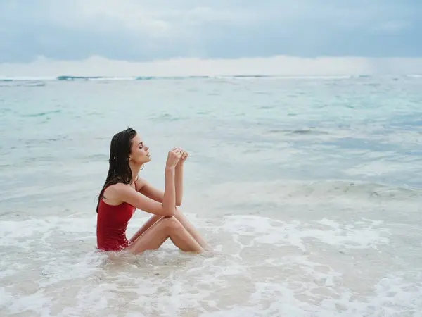 波の中の海岸の砂の上に座っている赤い水着の美しいタンツーリストと女性 高品質の写真 — ストック写真