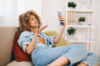 Rahat bir kadın koltukta oturuyor, cep telefonunu tutuyor, rahat oturma odasında kameraya gülümsüyor.