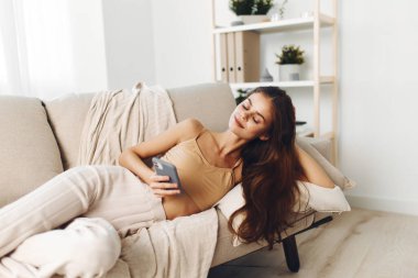 Cep telefonu tutan mutlu bir kadın, evdeki rahat koltukta dinleniyor, online oyun oynamanın tadını çıkarıyor..