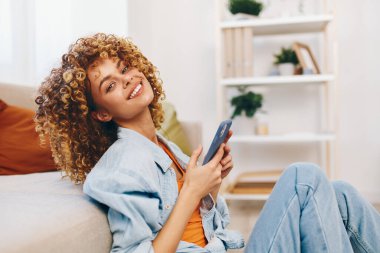 Neşeli kadın rahat koltukta oturuyor, cep telefonunu tutuyor ve oturma odasındaki kameraya gülümsüyor.