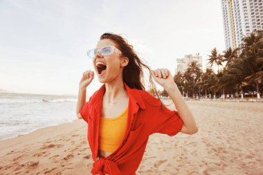 Gülümseyen Kadın Sahilde Özgürlüğün ve Renkli Günbatımının Tadını Çıkartıyor: Moda Güneş Gözlüklü Neşeli Kafkas Kız, Kaygısız Yaşam Tarzını Kucaklıyor