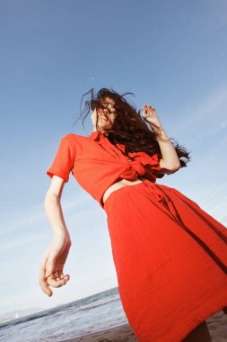 Güneşli plajda Neşeyle Dans eden Gülümseyen Kadın, Kırmızı Kıyafetlerle Özgürlüğün ve Eğlencenin tadını çıkartıyor
