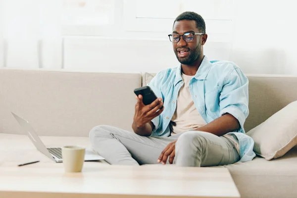Hymyilevä Afroamerikkalainen Mies Työskentelee Kannettavan Tietokoneen Kanssa Istuessaan Sohvalla Modernissa — kuvapankkivalokuva