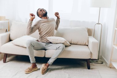 Rahat bir Afrikalı Amerikalı adam modern oturma odasında rahat bir koltukta oturuyor, gülümsüyor ve yastık tutuyor, evde huzurlu bir hafta sonu geçiriyor ve düşünüyor yalnız, teknolojisini kullanıyor.,