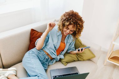 Modern oturma odasındaki rahat koltukta dizüstü bilgisayar kullanan gülümseyen kadın.