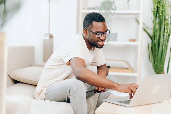 Smiling African American Freelancer Working on Laptop in Modern Home Office Young African American Guy Typing on Laptop and Relaxing on Sofa in Living Room Happy African American Student using Laptop