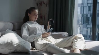 Evdeki kanepede oturan bir kadın telefonu elinde ve işe gitmeden önce bir kupadan içki içiyor. Yüksek kalite 4k görüntü