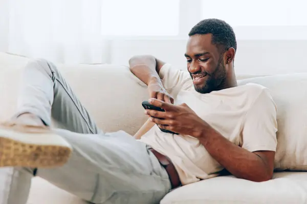 Щасливий Афроамериканський Чоловік Друкує Своєму Чорному Смартфоні Сидячи Сучасному Дивані — стокове фото