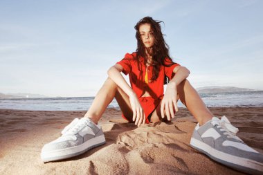 Neşeli bir kadın kumsalda oturuyor, tatilin tadını çıkarıyor ve şık bir yaz kıyafeti giyiyor..