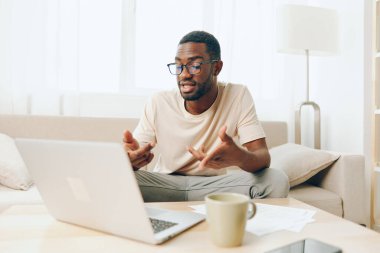 Gülümseyen Afrikalı Amerikalı Serbest Çalışan Modern Oturma Odası 'nda dizüstü bilgisayarında çalışan genç bir Afrikalı Amerikalı, rahat oturma odasındaki bir kanepede oturmuş bilgisayarında yazarken mutlu bir şekilde