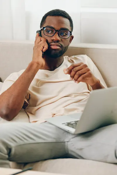 Усміхнений Афроамериканський Чоловік Сидить Дивані Використовуючи Ноутбук Роблячи Телефонний Дзвінок — стокове фото