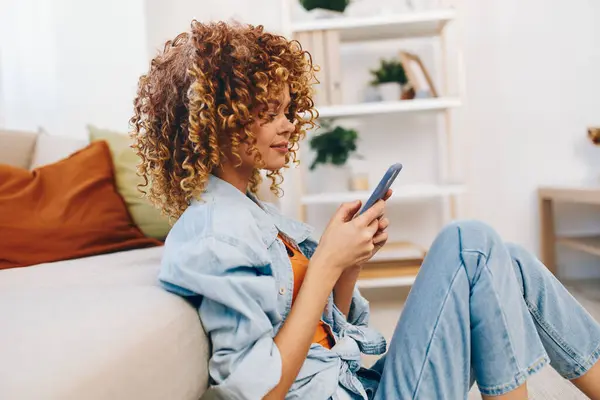 Happy Woman Holding Phone and Smiling, Enjoying Online Game on Sofa in Cozy Living Room