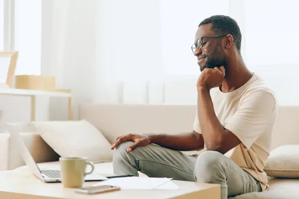 Black Millennial Freelancer Working on Laptop in Modern Home Office, Smiling and Typing African American guy sitting on a cozy sofa in his living room, enjoying the comforts of working from home With