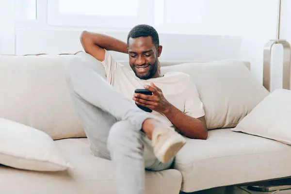 Happy African American man sitting on a black sofa, typing on his mobile phone and enjoying technology in the comfort of his modern apartment With a relaxed smile on his face, he effortlessly connects