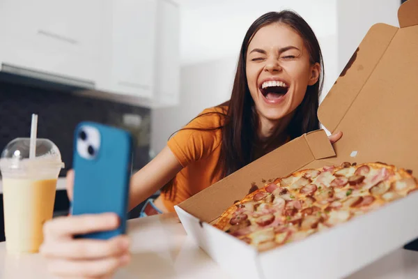 Woman taking a selfie with a slice of pizza in her hand and a pizza box in front of her