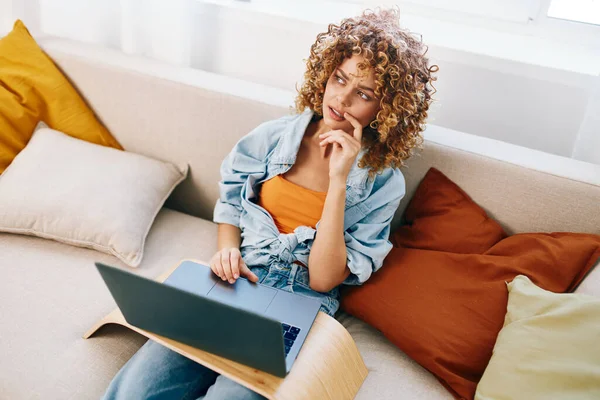 Evinden çalışan, dizüstü bilgisayarda yazan, kulaklık takan, modern bir oturma odasında rahat bir koltukta oturan gülümseyen bir kadın.