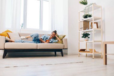 Evdeki rahat bir koltukta oturan rahat bir kadın cep telefonunu tutuyor ve internette sohbet ederken gülümsüyor..