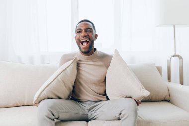 Rahatlamış Afrikalı Amerikalı adam, evinde modern bir koltukta oturuyor, ekstra rahatlık için bir yastıkla hafta sonu istirahatinin tadını çıkarıyor, gülümsüyor ve internetten bir şey izliyor, mutlu ve memnun bir şekilde oturma odasında oturuyor.
