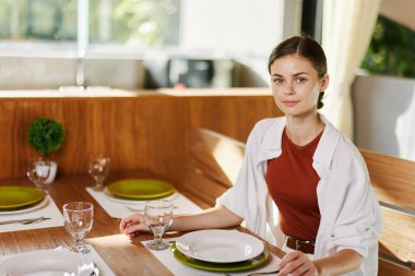 Şık bir kadın yemek masasında oturmuş bardaktan su içiyor, modern bir ev, gün batımı ışığı, rahat bir yaşam tarzı. Yüksek kalite fotoğraf