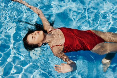 Havuzda yüzen kırmızı mayo giymiş, suda uzanan ve havuzda yüzen güzel bir gülümsemesi olan genç bir kadın. Tatilde dinlenme kavramı. Yüksek kalite fotoğraf