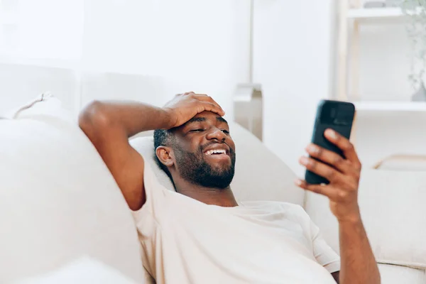 Happy African American man sitting on a black sofa, using his mobile phone He is casually dressed in a white Tshirt and is engrossed in reading and typing messages on his phone The modern apartment