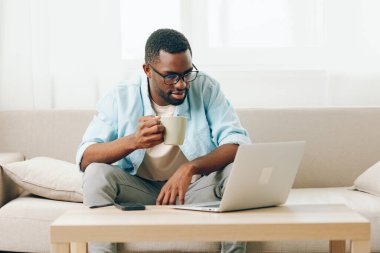 İçişleri Bakanlığı 'nda dizüstü bilgisayarda çalışan ve telefonda konuşan gülümseyen Afrikalı Amerikalı bir erkek, Afro-Amerikalı bir adam, rahat dairesinde kanepede oturuyor.