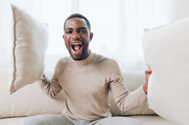 Gülümseyen Afrikalı Amerikalı bir adam modern oturma odasında rahat bir koltukta oturuyor, elinde bir yastıkla, rahat ve mutlu görünüyor, hafta sonunun keyfini çıkarıyor diye düşünüyor.