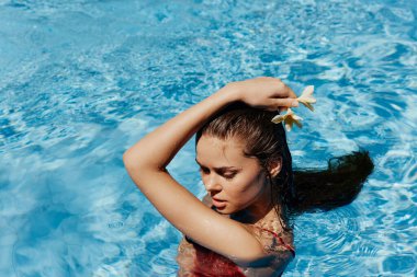 Yüzme havuzunda yüzen, kırmızı mayolu, güzel gülüşlü ve havuzda yüzen Bali çiçekli genç bir kadın. Tatilde rahatlama kavramı. Yüksek kalite fotoğraf