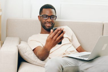 Gülümseyen Afrikalı Amerikalı Adam Laptop 'ta çalışıyor ve Konforlu Oturma odasında telefonla konuşuyor modern serbest çalışan kişi dairesindeki rahat bir kanepede oturuyor, gözlük takıyor ve daktilo yazıyor.
