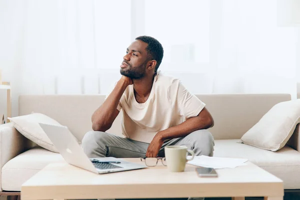 Sad African American Freelancer Working on Laptop in a Modern Home In this image, a tired and unhappy African American freelancer sits on a sofa in a stylish living room He is working on his laptop,