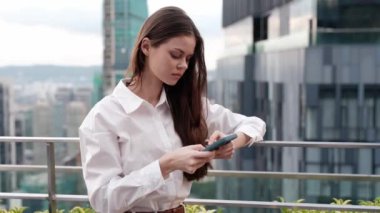 Elinde telefonu olan bir iş kadını büyük şehir gökdelenleri ve iş merkezinin arka planındaki bir binanın çatısına mesaj yazıyor. İş seyahati. Yüksek kalite 4k görüntü