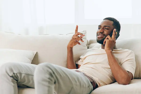 Happy African American Man Talking on a Black Sofa, Using a Smartphone for a Video Call at Home This image depicts a young, confident African American man sitting on a modern black sofa, engaged in a
