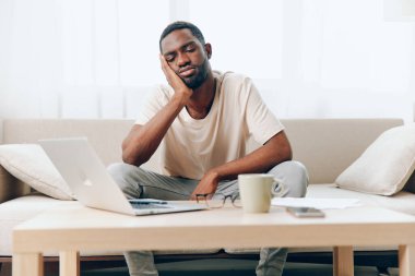 Modern bir ofiste dizüstü bilgisayar üzerinde çalışan yorgun Afrikalı Amerikalı bir adam bir koltukta oturuyor, üzüntü ve mutsuzluğu ifade ediyor, muhtemelen bir krizle karşı karşıya.