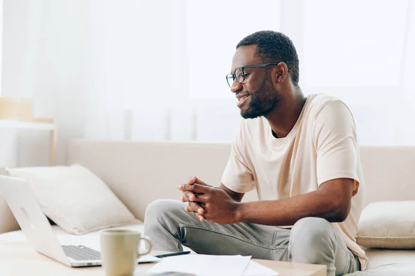 Smiling African American Freelancer Working on Laptop in Modern Home Office A young African American man with a casual style is sitting on a comfortable sofa in a modern living room He is happily