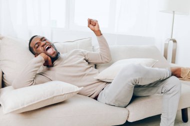 Rahatlamış Afrikalı Amerikalı Adam, evinde koltukta oturuyor, dinlendirici bir hafta sonunun tadını çıkarıyor, gülümsüyor ve yastık tutuyor, o, Modern Oturma Odasının rahatlığında, Mutluluk ve Mutluluk Arkaplanı ile.