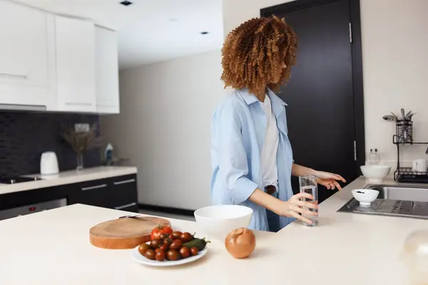 stock image A woman standing in a kitchen with a bowl of fruit and a glass of water, healthy lifestyle concept