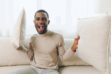 Rahat, Afro-Amerikan bir adam, rahat ve şık oturma odasında, arkasında bir yastıkla, evinde hafta sonunun tadını çıkarırken gülümsüyor.