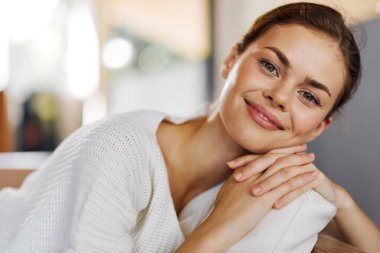 Mutlu bir kadın, evinde rahat bir koltukta oturuyor, gevşiyor ve gülümsüyor, neşeli bir gülümsemeyle, rahat bir süveter giyiyor, sıcak ve çekici bir oturma odasında dinleniyor.