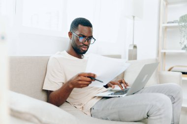 Gülümseyen Afrikalı Amerikalı Serbest Yazıcı, bu modern ve rahat oturma odasında çalışırken, genç Afrikalı Amerikalı bir adam kanepede oturuyor, dizüstü bilgisayarıyla çalışıyor.