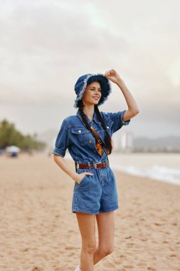 Yaz Rüzgarındaki Güzellik: Sahilde Rahatlayan Çekici Genç Kadın, Beyaz Şapka ve Mavi Çiçek Elbisesiyle Deniz ve Gökyüzünün tadını Çıkartıyor