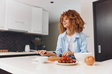 Kahvaltının tadını çıkaran kıvırcık saçlı kadın mutfak masasında bir tabak yemek ve bir fincan kahveyle oturuyor.