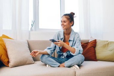 Mutlu kadın, evinde rahat bir koltukta oturuyor, cep telefonunu tutuyor ve çevrimiçi bir oyun oynarken gülümsüyor..